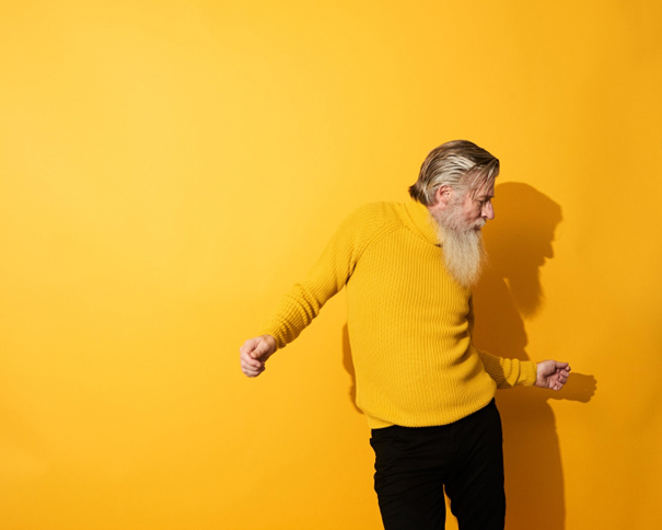 Ein eldre mann med sjegg som dansar framfor ein gul vegg