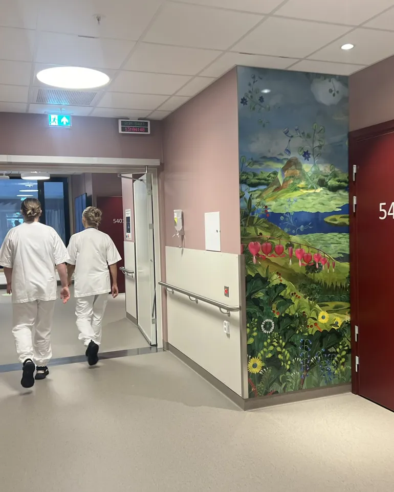 Et par sykepleiere som går gjennom en gang med et stort maleri på veggen