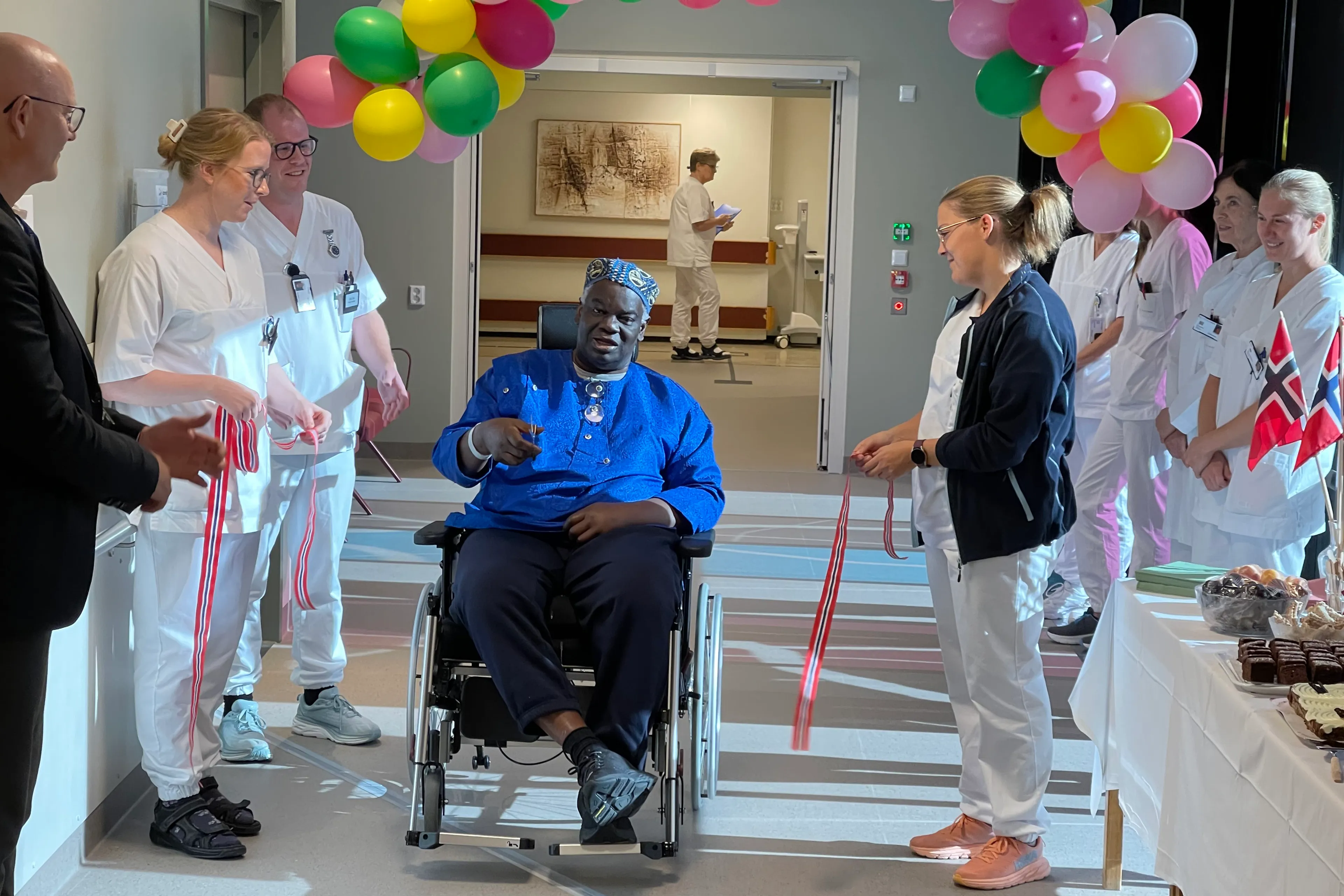 Ein mann i rullestol og ei rekke helsepersonell i ein gang med ballongar og flagg