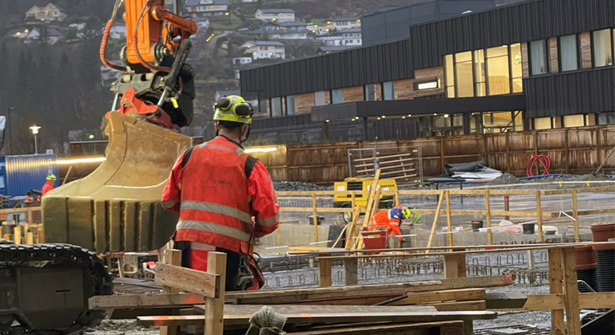 Ein mann iført arbeidskle jobbar på ein byggeplass. I bakgrunnen ein grabb frå ei gravemaskin og delar av eit bygg