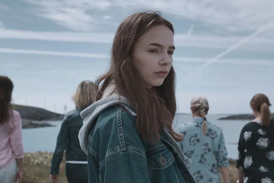 Ei ung jente med dongerijakke som står ute, med fleire ungdomar i bakgrunnen