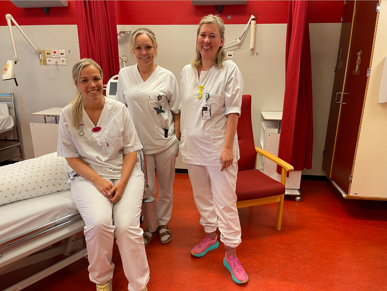 tre kvinnelege sjukepleiarar som er kledd i kvit uniform inne på eit pasientrom med raude gardiner og møblement.